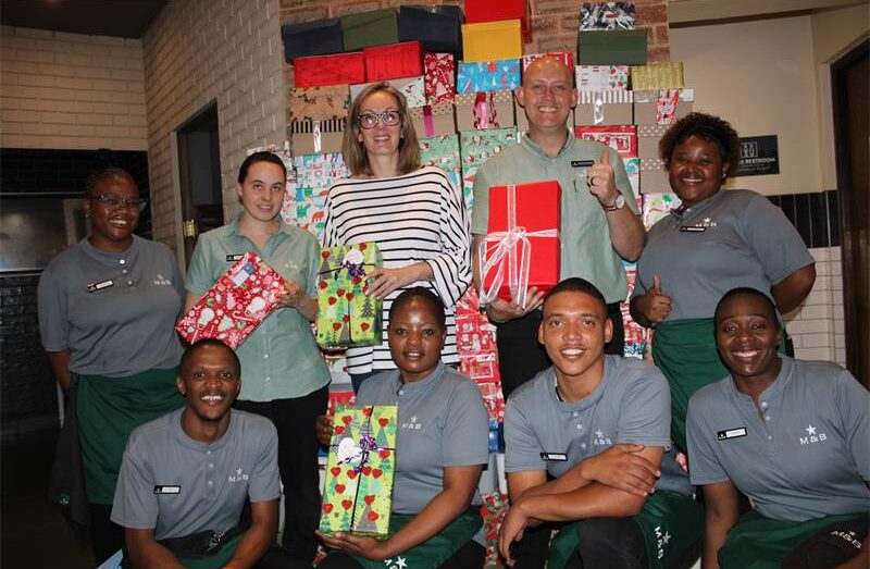 Group of people standing in front of a stack of Christmas gifts with some holding boxes in their hands