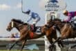 Two jockeys racing brown horses with a woman in the background taking photos.