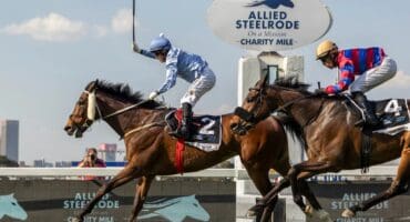 Two jockeys racing brown horses with a woman in the background taking photos.