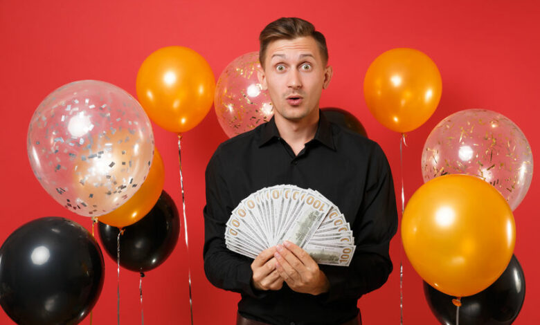 Shocked amazed young man in classic shirt holding bundle lots of dollars, cash money on red background air balloons. International Women's Day, Happy New Year, birthday mockup holiday party concept.