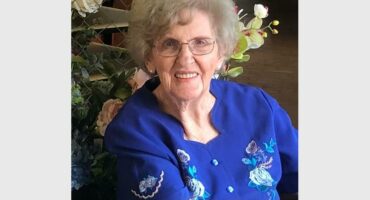 An older woman in blue shirt sitting in front of a flower arrangement.