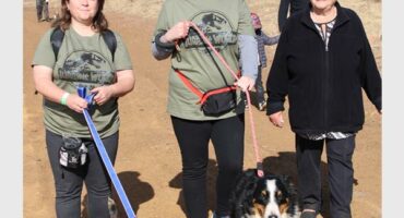 Three women and two dogs walking on a dirt road.