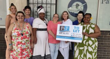 Joyful real estate agents and community members holding a "To Let" sign outside Brakpan Herald office.