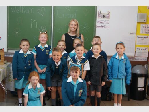 Graad 1 'n Groepie van 11 wit kinders in turkoois en donker turkoois skool uniforms bondel om 'n volwasse wit vrou in 'n swart top saam.