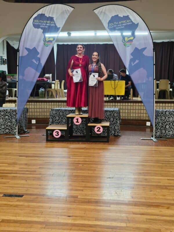 Two girls on the first and second place spots on a podium, both wearing long maroon flowy dresses and holding certificates, with medals around their necks.