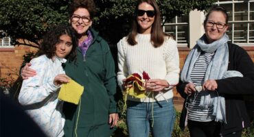 Four women are standing together holding knitting needles.