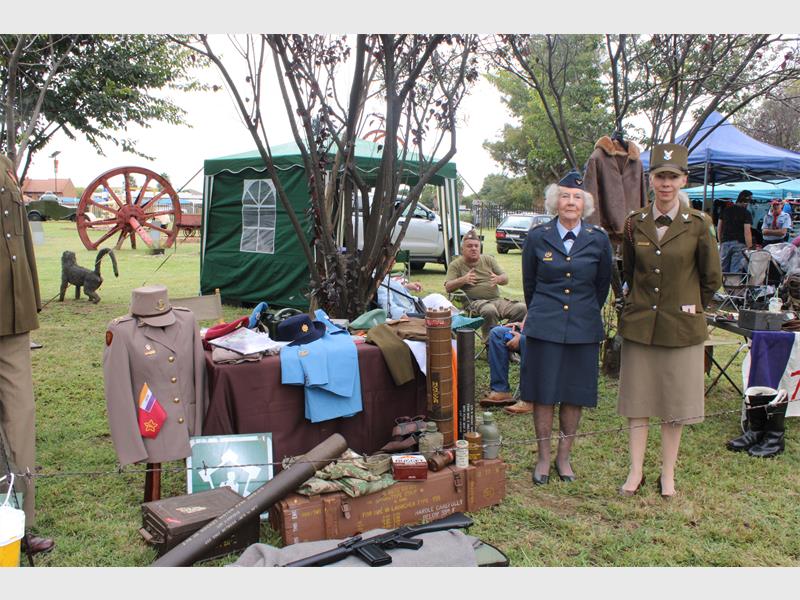 Two women in vintage military uniforms stand beside a display of historical military memorabilia, including uniforms, hats, and old equipment. The scene conveys a sense of nostalgia and historical appreciation.