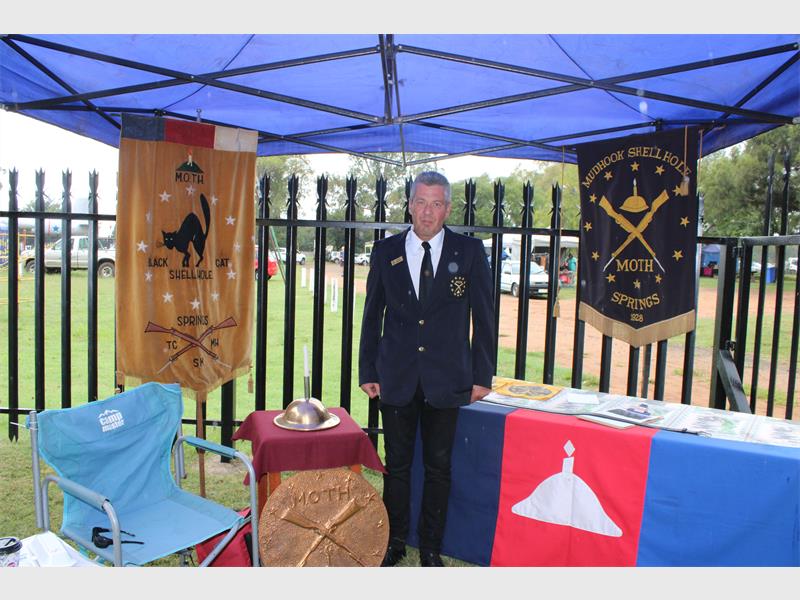 A man in a formal blazer stands under a blue canopy. He's surrounded by military memorabilia, banners, and a table with brochures, conveying respect and remembrance.