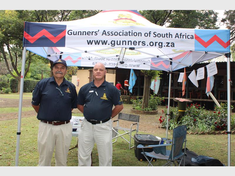 Two men stand under a tent labeled "Gunners' Association of South Africa." They wear matching navy shirts with logo patches, conveying a professional and welcoming tone.