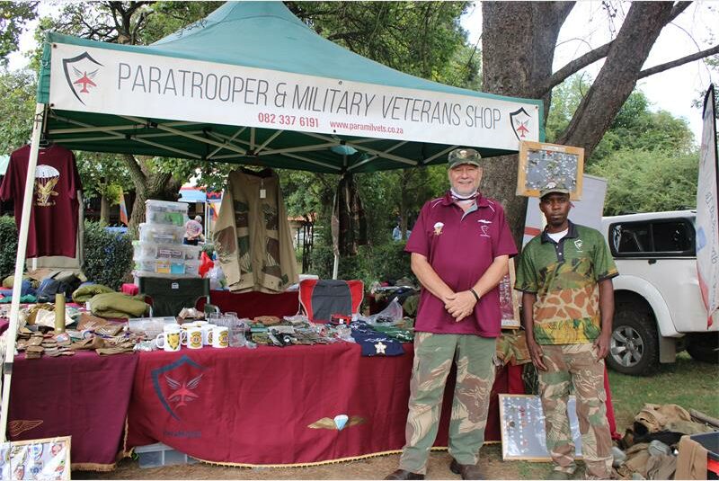 Two men in camouflage pants stand in front of a tent labeled "Paratrooper & Military Veterans Shop" with military-themed items on display.