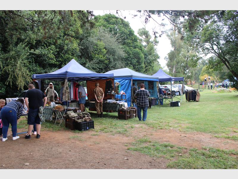 Outdoor market scene with people browsing under blue tents. Stalls display clothing and other items. The area is surrounded by lush greenery, creating a relaxed, community atmosphere.