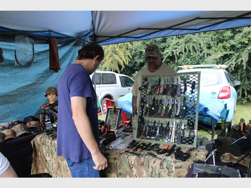 A man browses a market stall displaying various knives on a camo-patterned tablecloth under a blue canopy. Hats and other items are also displayed. The scene is outdoors with a car and greenery in the background, creating a casual, rugged atmosphere.