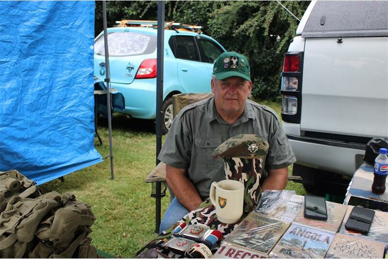 A man in a green hat sits at an outdoor table displaying military memorabilia, including badges and books. A blue car and a white truck are in the background.