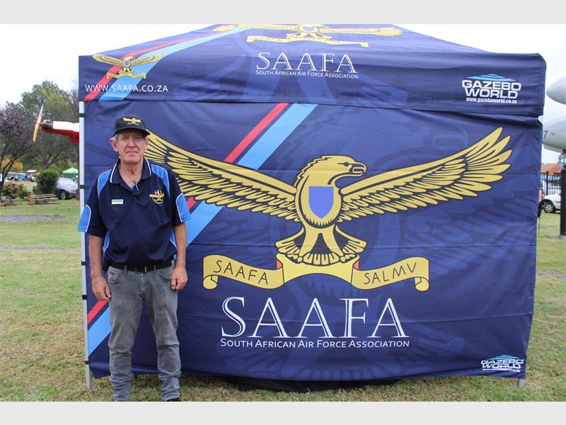 A man stands in front of a blue tent featuring the South African Air Force Association logo, with a large eagle graphic. He wears a SAAFA hat and shirt.