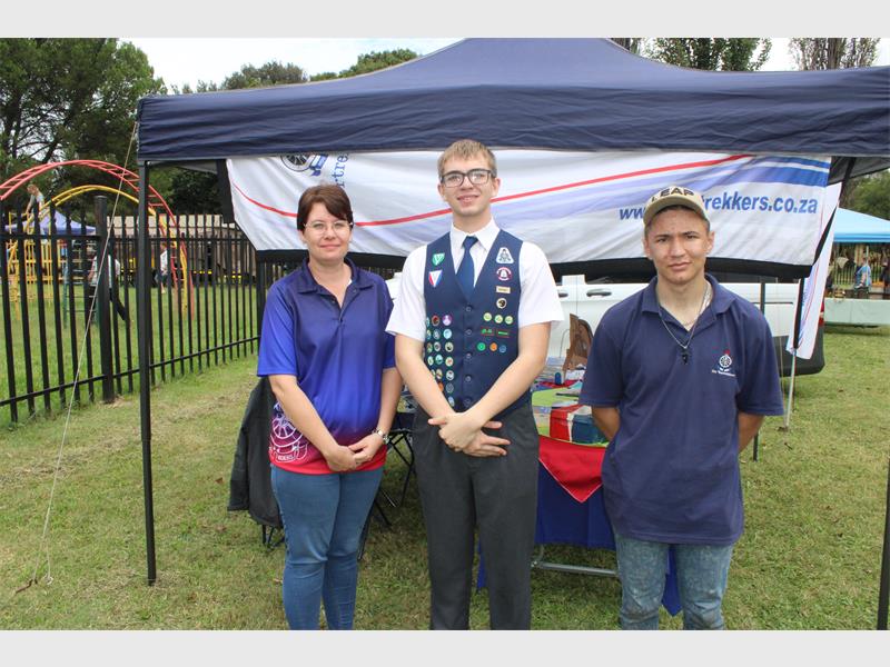 Three individuals stand in front of a "Voortrekkers" tent at an outdoor event. The central figure wears a badge-covered vest, with a relaxed and cheerful atmosphere.