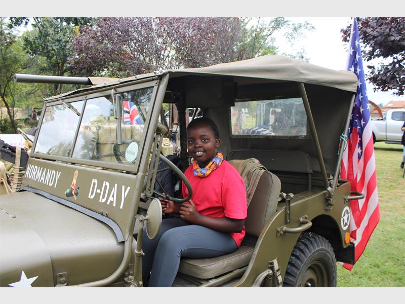 A smiling child in a pink shirt sits in a vintage military jeep labeled "Normandy D-Day." The jeep, adorned with American flags, is parked outdoors amidst trees, creating a cheerful, historical atmosphere.