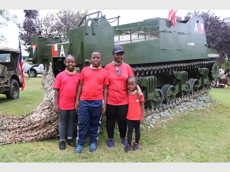A family of four, all in red shirts, stands smiling in front of a large green military vehicle on grass, conveying a sense of unity and pride.