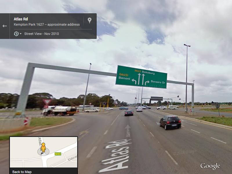 Street view of Atlas Road, showing a busy multi-lane intersection with directional signage indicating routes to Brenner and Boksburg. Cloudy sky overhead.
