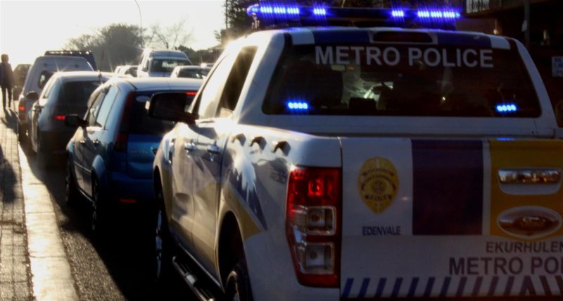A white Metro Police vehicle with flashing blue lights is parked next to a blue car on a busy road, creating a tense atmosphere in the morning light.