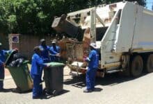 Sanitation workers in blue uniforms collect trash bins into a white garbage truck on a sunny day. Teamwork and efficiency are evident.