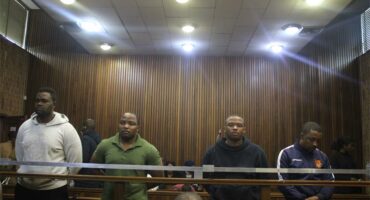Four men stand in a courtroom behind a wooden barrier, with serious expressions. The room features wooden paneling and overhead lights, conveying a formal atmosphere.