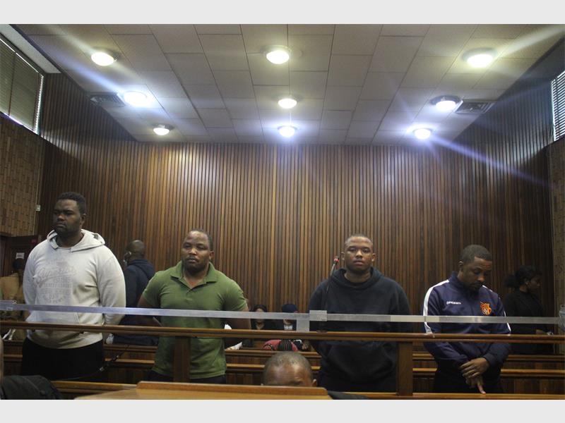 Four men stand in a courtroom behind a wooden barrier, with serious expressions. The room features wooden paneling and overhead lights, conveying a formal atmosphere.