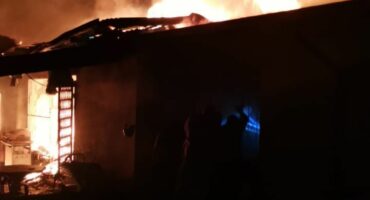 Building engulfed in flames at night, with intense orange and yellow fire illuminating the scene. Dark smoke rises above, creating a dramatic, urgent atmosphere.