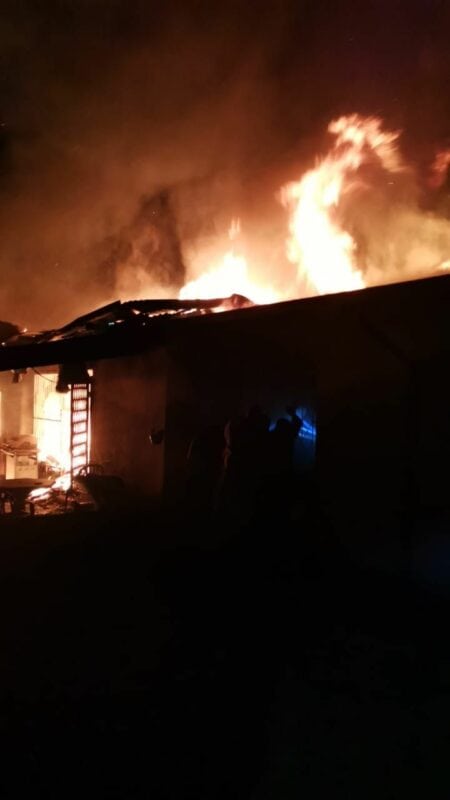 Building engulfed in flames at night, with intense orange and yellow fire illuminating the scene. Dark smoke rises above, creating a dramatic, urgent atmosphere.