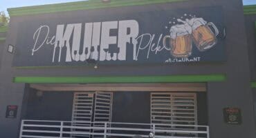 Sign for "Die Kuier Plek" restaurant with two frothy beer mugs. Modern gray building with green trim, white railing, and a motorcycle outside.