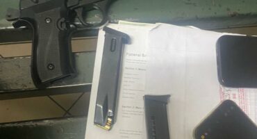 A black semi-automatic pistol beside two magazines, paperwork, and mobile phones on a green surface.