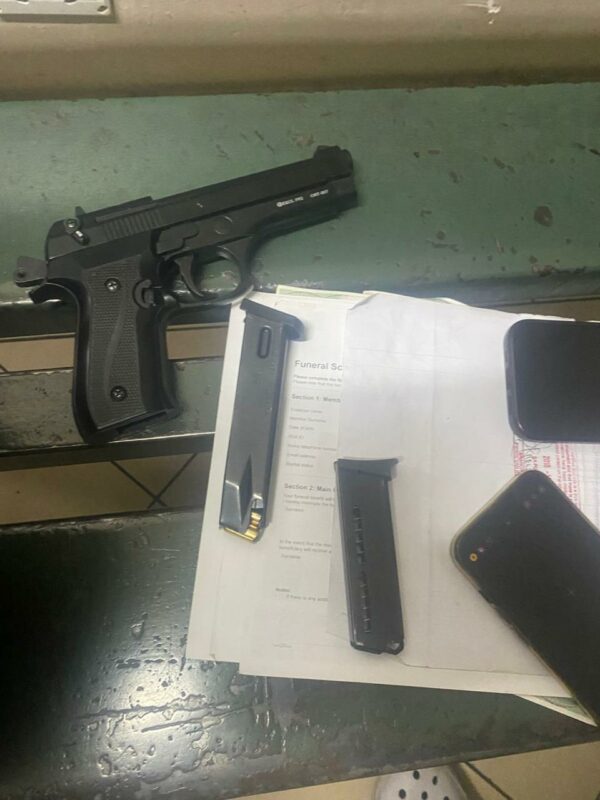 A black semi-automatic pistol beside two magazines, paperwork, and mobile phones on a green surface.
