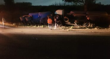 Two trucks are overturned on a dimly lit roadside at night. Three people, one in a reflective jacket, stand nearby, suggesting a recent accident scene