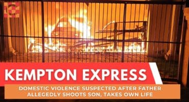 A car engulfed in bright, intense flames behind a metal gate at night. The scene suggests urgency and danger as smoke rises into the dark sky.