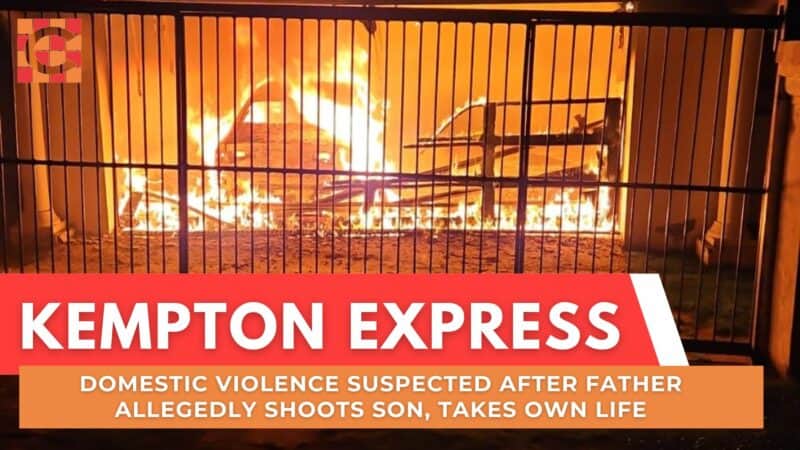 A car engulfed in bright, intense flames behind a metal gate at night. The scene suggests urgency and danger as smoke rises into the dark sky.