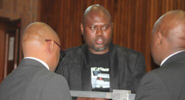Two men in suits are engaged in conversation with Thabani Goodwill Ntshalintshali in a courtroom setting, with wooden paneling visible in the background.