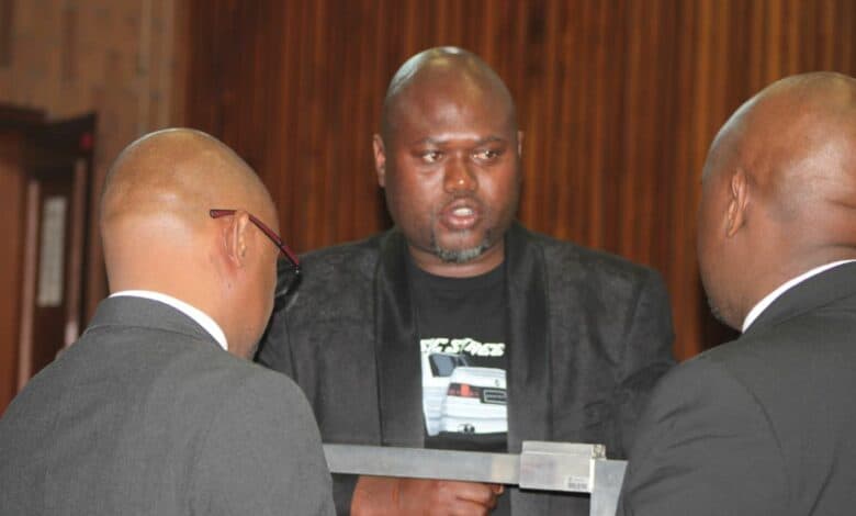 Two men in suits are engaged in conversation with Thabani Goodwill Ntshalintshali in a courtroom setting, with wooden paneling visible in the background.