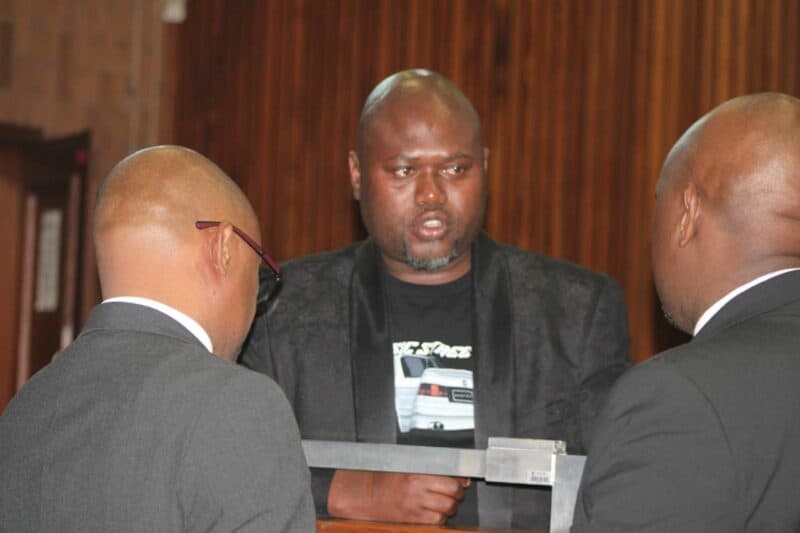 Two men in suits are engaged in conversation with Thabani Goodwill Ntshalintshali in a courtroom setting, with wooden paneling visible in the background.