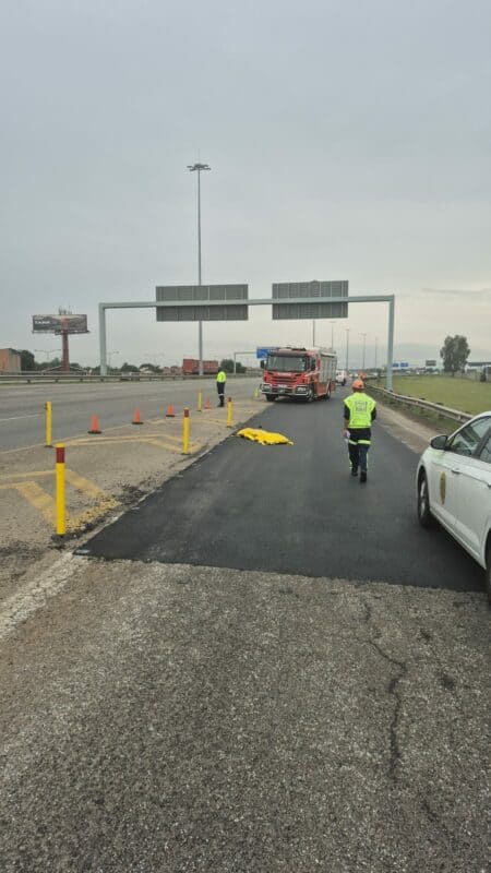 Emergency personnel manage a scene on a highway with a fire truck, traffic cones, and a covered object. Overcast sky and a calm, controlled atmosphere.