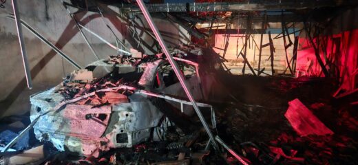 Burned-out car amidst debris in a charred and collapsed garage. Dim lighting with red hues creates a somber and devastating atmosphere.