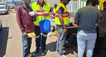 Two men in safety vests interact with others on a sunny street. One holds a blue bag and pamphlets, smiling. A colorful umbrella shades a vendor table.