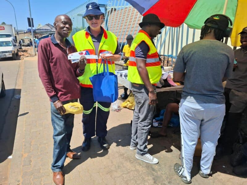 Two men in safety vests interact with others on a sunny street. One holds a blue bag and pamphlets, smiling. A colorful umbrella shades a vendor table.