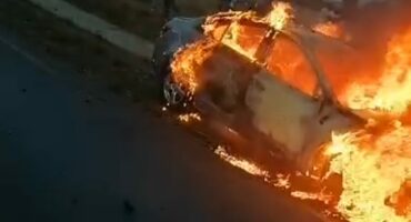 A car is engulfed in bright orange flames on a roadside, with dark smoke billowing. Passersby are visible in the background, creating a tense scene.