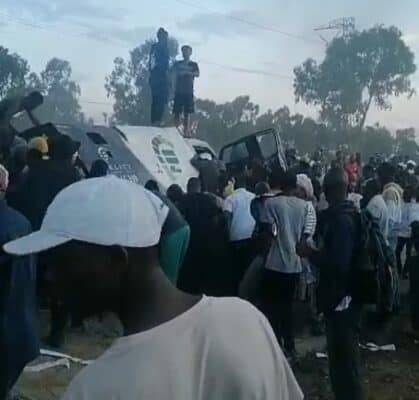 A crowd gathers around and on top of an overturned bus, looking concerned. Trees and faint structures fill the background, creating a tense atmosphere.