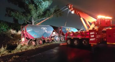 A large tow truck with bright lights is pulling a damaged trailer off an embankment at night. Wet road reflects light, creating a dramatic scene.