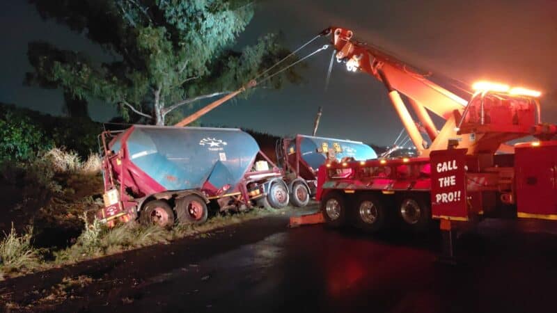 A large tow truck with bright lights is pulling a damaged trailer off an embankment at night. Wet road reflects light, creating a dramatic scene.