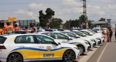 A line of white patrol cars with blue and yellow EMPD markings are parked on the roadside. Officers in orange vests stand nearby under a cloudy sky.