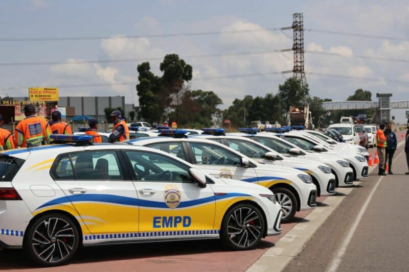 A line of white patrol cars with blue and yellow EMPD markings are parked on the roadside. Officers in orange vests stand nearby under a cloudy sky.