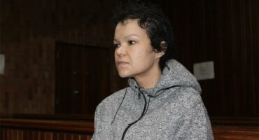 Denise Hattingh with short curly hair sits in a wooden courtroom wearing a gray hoodie. The atmosphere is tense and solemn, with dim lighting enhancing the serious mood.