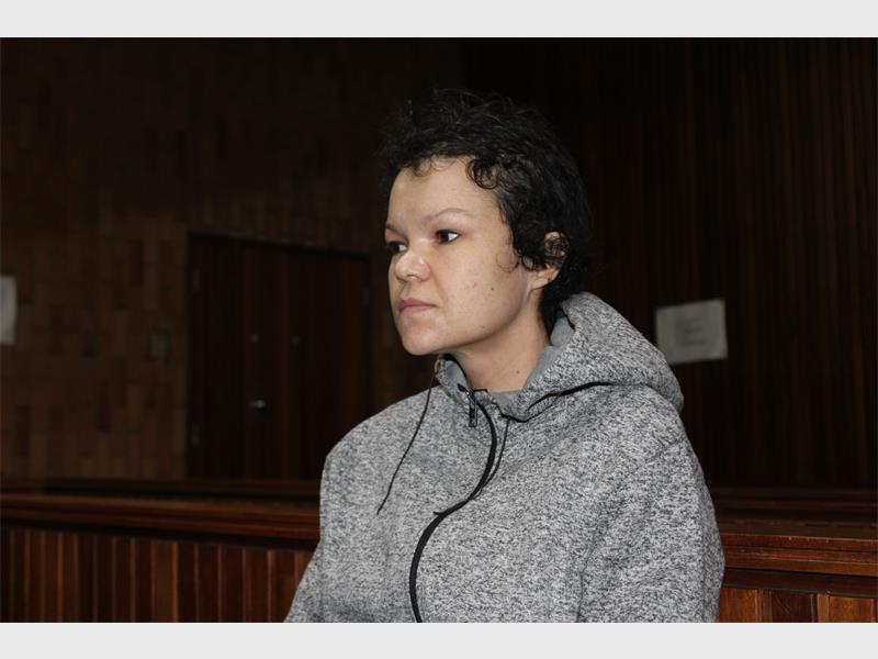 Denise Hattingh with short curly hair sits in a wooden courtroom wearing a gray hoodie. The atmosphere is tense and solemn, with dim lighting enhancing the serious mood.