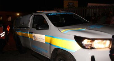 A white police vehicle with bright blue and yellow stripes is parked at night, its headlights are on. People and buildings are dimly visible in the background.